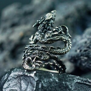 May include: A detailed silver dragon ring. The ring features a dragon wrapped around the finger, with intricate scales and a detailed head. The ring is set against a dark, textured background, highlighting the ring's design.
