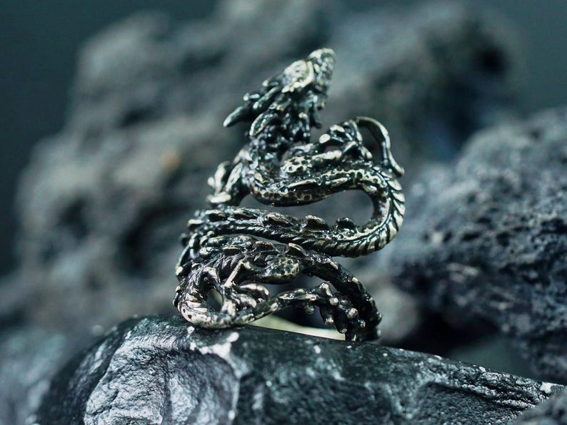 May include: A detailed silver dragon ring. The ring features a dragon wrapped around the finger, with intricate scales and a detailed head. The ring is set against a dark, textured background, highlighting the ring's design.