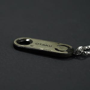 May include: A silver chain necklace with a silver pendant that says "OTAKU" on it. The pendant is oval shaped and has a small hole in the top for the chain to go through.