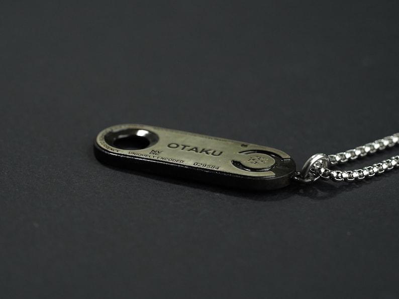 May include: A silver chain necklace with a silver pendant that says "OTAKU" on it. The pendant is oval shaped and has a small hole in the top for the chain to go through.