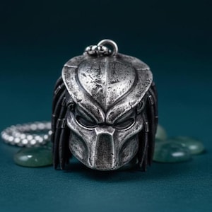 May include: A silver-colored pendant in the shape of a Predator mask. The pendant has intricate details, including the mask's distinctive features and dreadlocks. The pendant is attached to a silver chain.