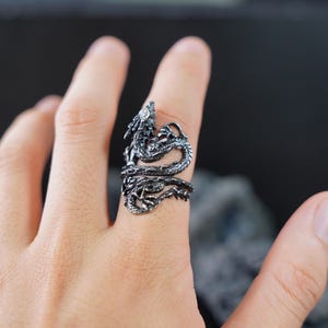 May include: A detailed, silver-colored dragon ring wraps around a finger. The dragon's head is raised, and its body coils around the finger. The ring has intricate detailing, showcasing scales and other features.