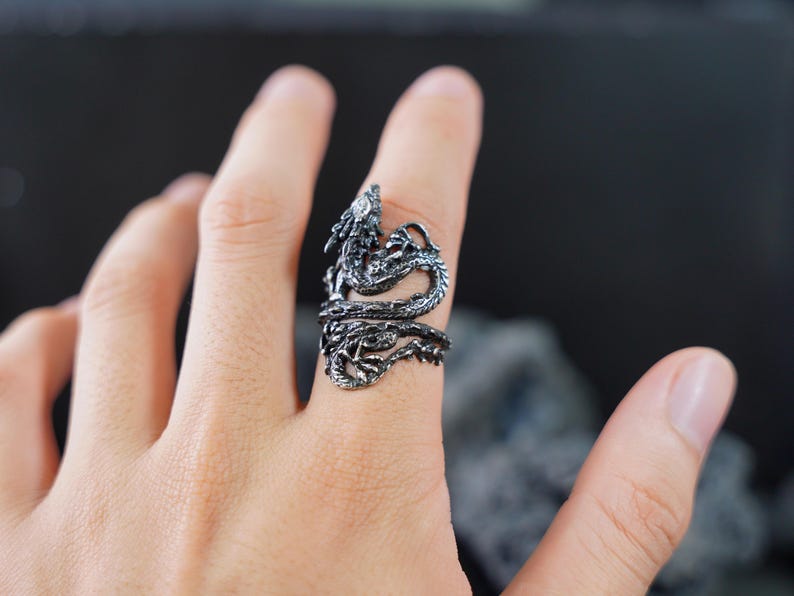 May include: A detailed, silver-colored dragon ring wraps around a finger. The dragon's head is raised, and its body coils around the finger. The ring has intricate detailing, showcasing scales and other features.