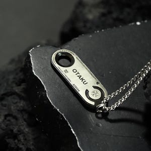 May include: A silver necklace with a rectangular pendant that says "OTAKU" and has a unique design. The pendant is engraved with the words "uniquely encoded" and a number.