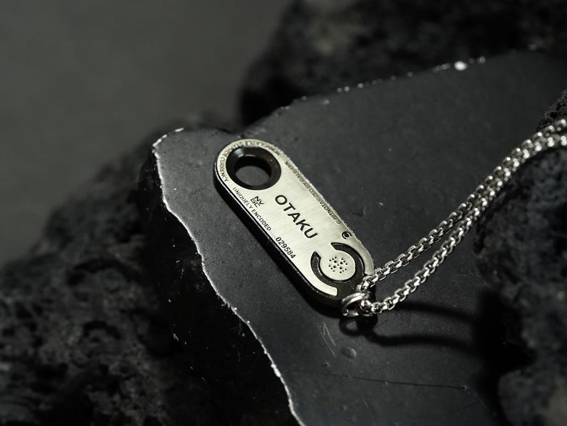 May include: A silver necklace with a rectangular pendant that says "OTAKU" and has a unique design. The pendant is engraved with the words "uniquely encoded" and a number.