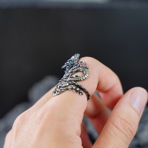 May include: A detailed silver dragon ring with intricate scales and a serpentine design. The dragon wraps around the finger, with its head and tail meeting. The ring is worn on a finger, showcasing the detailed craftsmanship.