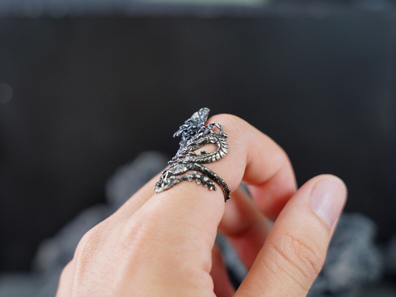 May include: A detailed silver dragon ring with intricate scales and a serpentine design. The dragon wraps around the finger, with its head and tail meeting. The ring is worn on a finger, showcasing the detailed craftsmanship.