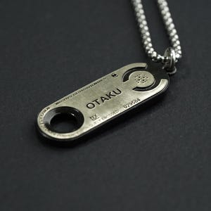 May include: A silver necklace with a pendant that says "OTAKU" and has a circular design. The pendant is attached to a silver chain.