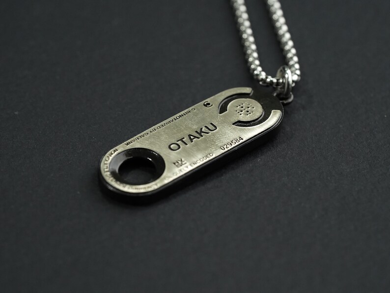 May include: A silver necklace with a pendant that says "OTAKU" and has a circular design. The pendant is attached to a silver chain.