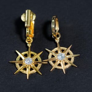 K&ouml;nnte beinhalten: Ein Paar goldfarbene Ohrclips mit einem Sternendesign und einem klaren Strassstein in der Mitte.