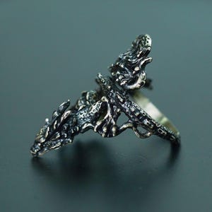 May include: A detailed silver dragon ring. The ring features a dragon design with intricate scales and a textured surface. The dragon's head and body wrap around the finger, creating a unique and eye-catching accessory.