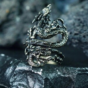 May include: A detailed silver-colored dragon ring. The dragon wraps around the finger, with intricate scales and a detailed head. The ring is set against a dark, textured background, highlighting the ring's design.