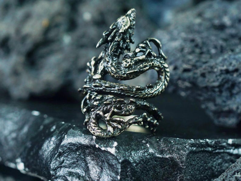 May include: A detailed silver-colored dragon ring. The dragon wraps around the finger, with intricate scales and a detailed head. The ring is set against a dark, textured background, highlighting the ring's design.