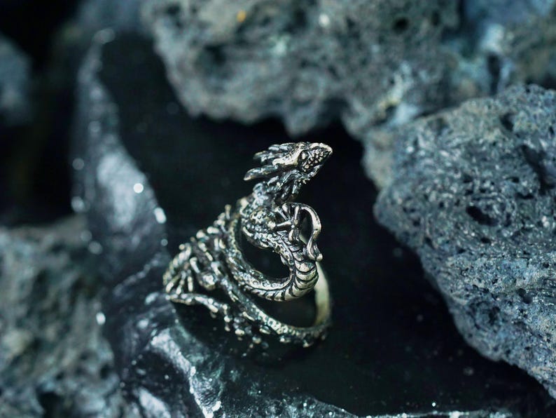 May include: A silver-colored dragon ring with intricate detailing. The ring features a dragon design with its head raised and mouth open. The ring is set against a dark, textured background.