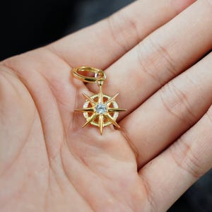 K&ouml;nnte beinhalten: Ein goldener Kompass-Anh&auml;nger mit einem klaren Kristall in der Mitte. Der Kompass hat acht Punkte und ist an einem goldenen Ring befestigt.