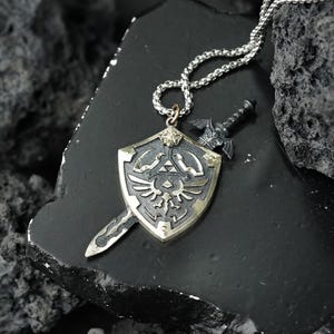 Può includere: Una collana con catena d'argento con un ciondolo che presenta una spada d'argento e uno scudo dorato con un designo alato. Lo scudo ha un simbolo triangolare con un triangolo più piccolo all'interno.