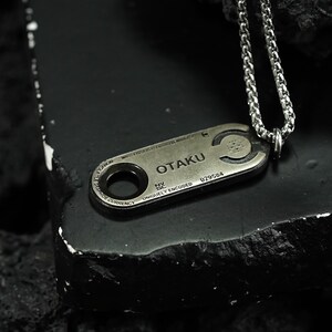 May include: A silver chain necklace with a silver pendant that says "OTAKU" and has a unique code. The pendant is shaped like a small rectangular tag with a circular hole in the center.