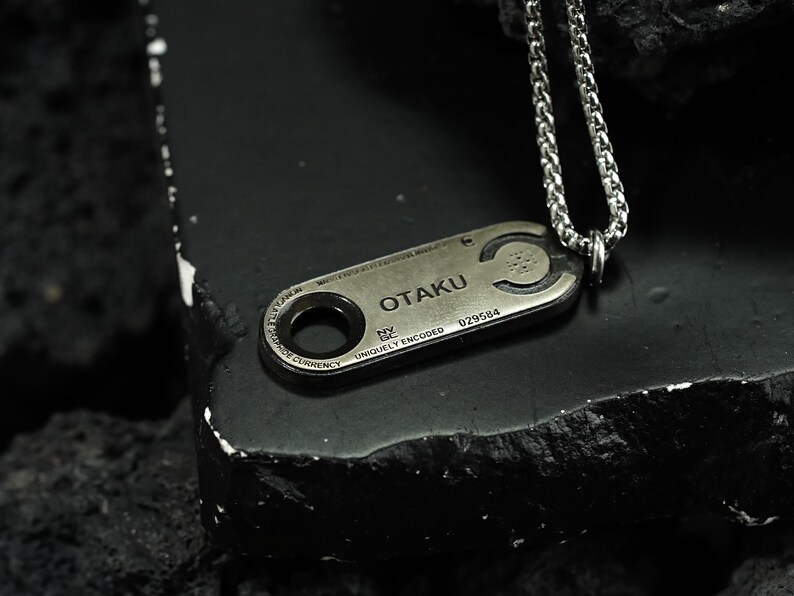 May include: A silver chain necklace with a silver pendant that says "OTAKU" and has a unique code. The pendant is shaped like a small rectangular tag with a circular hole in the center.
