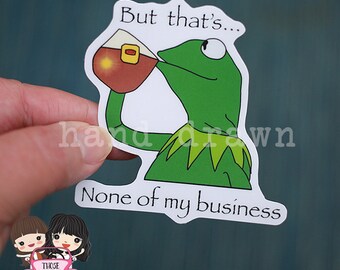 May include: A white sticker with a green frog drinking from a red cup. The sticker says "But that's... None of my business".