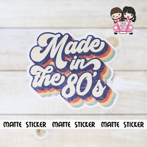 May include: A matte sticker with the text "Made in the 80's" in a retro font, featuring a blue, red, and yellow color scheme. The sticker has a distressed look and is surrounded by a rainbow-colored border. The words "Matte Sticker" are printed at the bottom.