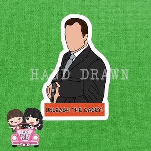 Puede incluir: Una pegatina con una ilustración de dibujos animados de un hombre con un traje negro sosteniendo una pistola, con el texto "UNLEASH THE CASEY!" en una pancarta roja. El fondo es verde y la pegatina tiene un borde blanco. También son visibles las palabras "HAND DRAWN".
