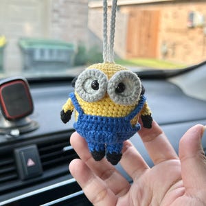 Może przedstawiać: Szydełkowy urok samochodu Minion. Urok ma ż&oacute;łte ciało, niebieskie ogrodniczki i duże oczy z szarymi obw&oacute;dkami. Czarne detale obejmują buty i ramiona. Do zawieszenia przymocowany jest biały sznurek.