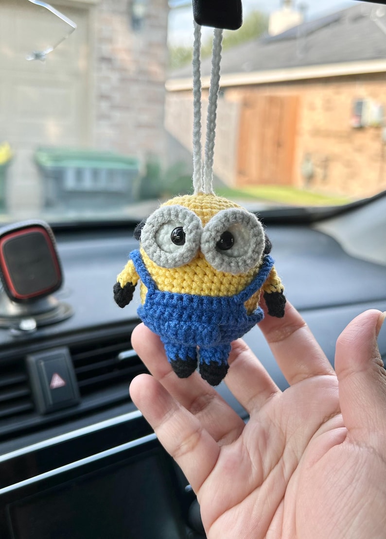 Może przedstawiać: Szydełkowy urok samochodu Minion. Urok ma ż&oacute;łte ciało, niebieskie ogrodniczki i duże oczy z szarymi obw&oacute;dkami. Czarne detale obejmują buty i ramiona. Do zawieszenia przymocowany jest biały sznurek.