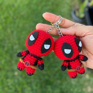 May include: Two red and black crochet Deadpool keychains. Each keychain features a round head with white eyes and a black mask, a red body, and black arms and legs. The keychains are connected by a silver chain.