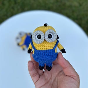 Może przedstawiać: Szydełkowa figurka Minionka, z ż&oacute;łtym korpusem, niebieskimi ogrodniczkami i dużymi oczami z szarymi obw&oacute;dkami. Ręcznie robiona zabawka ma czarne akcenty na dłoniach i stopach. Drugi, częściowo widoczny Minionek jest w tle.