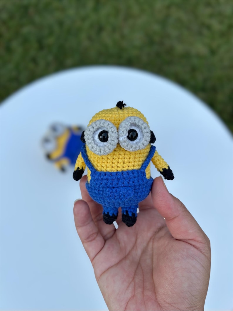 Może przedstawiać: Szydełkowa figurka Minionka, z ż&oacute;łtym korpusem, niebieskimi ogrodniczkami i dużymi oczami z szarymi obw&oacute;dkami. Ręcznie robiona zabawka ma czarne akcenty na dłoniach i stopach. Drugi, częściowo widoczny Minionek jest w tle.