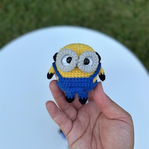 Może przedstawiać: Ręcznie robiona szydełkowa figurka Minionka, z ż&oacute;łtym korpusem, niebieskimi ogrodniczkami i dużymi szarymi oczami. Figurka ma czarne akcenty i jest trzymana w dłoni. Zabawka jest kapryśnym przedstawieniem popularnej postaci z kresk&oacute;wek.
