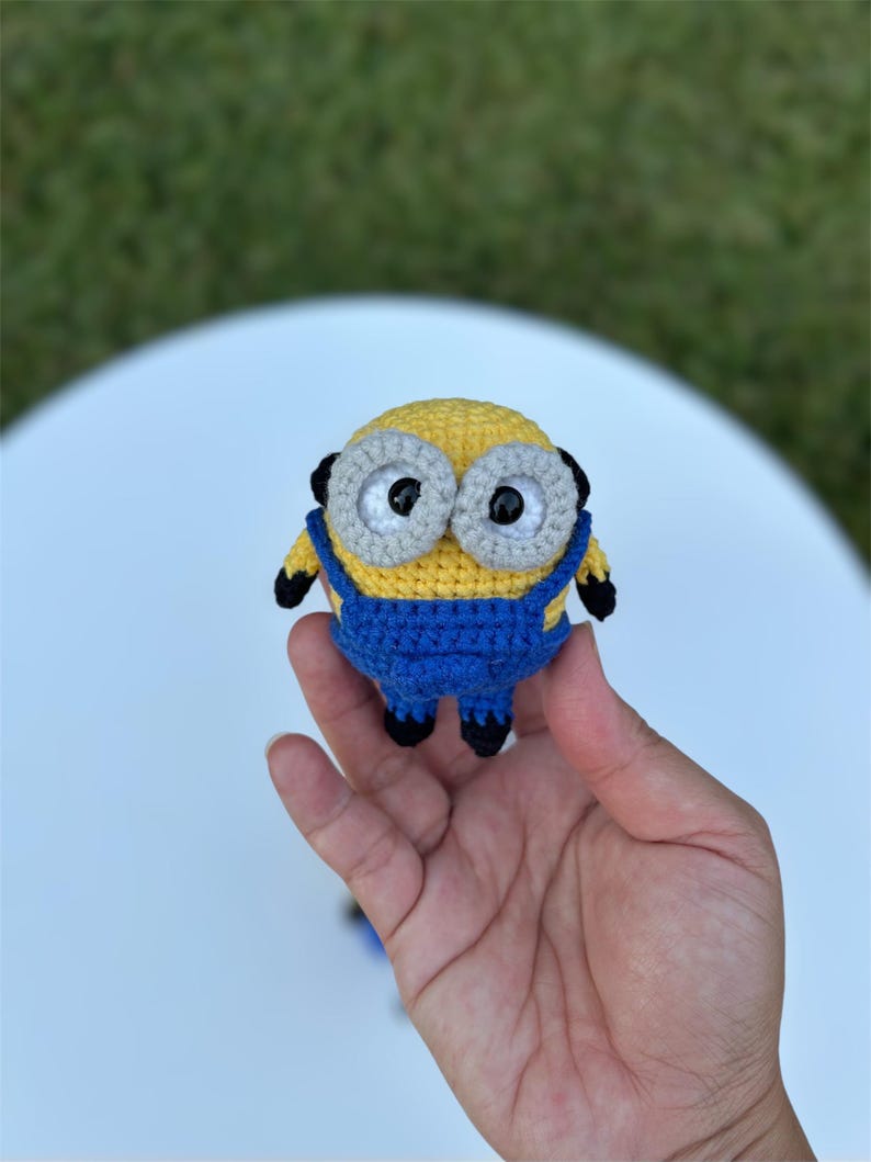 Może przedstawiać: Ręcznie robiona szydełkowa figurka Minionka, z ż&oacute;łtym korpusem, niebieskimi ogrodniczkami i dużymi szarymi oczami. Figurka ma czarne akcenty i jest trzymana w dłoni. Zabawka jest kapryśnym przedstawieniem popularnej postaci z kresk&oacute;wek.
