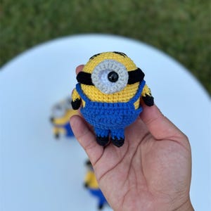 Może przedstawiać: Szydełkowa figurka Minionka w kolorze ż&oacute;łtym i niebieskim, z czarnymi akcentami. Figurka ma jedno oko i nosi niebieskie ogrodniczki. Figurka ma około 7,6 cm wysokości.