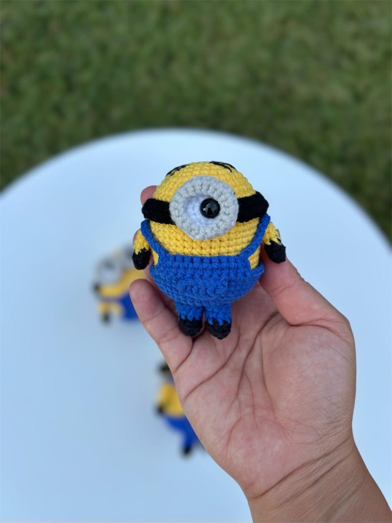 Może przedstawiać: Szydełkowa figurka Minionka w kolorze ż&oacute;łtym i niebieskim, z czarnymi akcentami. Figurka ma jedno oko i nosi niebieskie ogrodniczki. Figurka ma około 7,6 cm wysokości.