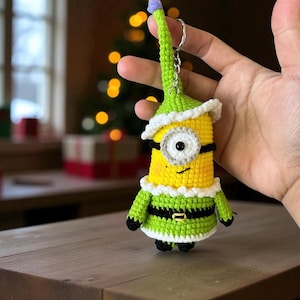 Może przedstawiać: Ręcznie robiony brelok z szydełka przedstawiający ż&oacute;łtą postać Minionka ubraną w zielony str&oacute;j elfa z białym kołnierzem i czapką. Brelok ma srebrny pierścień i fioletowy pompon na czapce. W tle widoczna jest choinka.