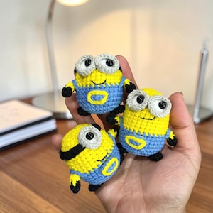 May include: Three small, handmade crochet figures inspired by Minions, featuring bright yellow bodies, blue overalls, and large white-rimmed eyes with black pupils. Each figure is crafted with detailed yarn stitching, showcasing a round shape and a cheerful expression.