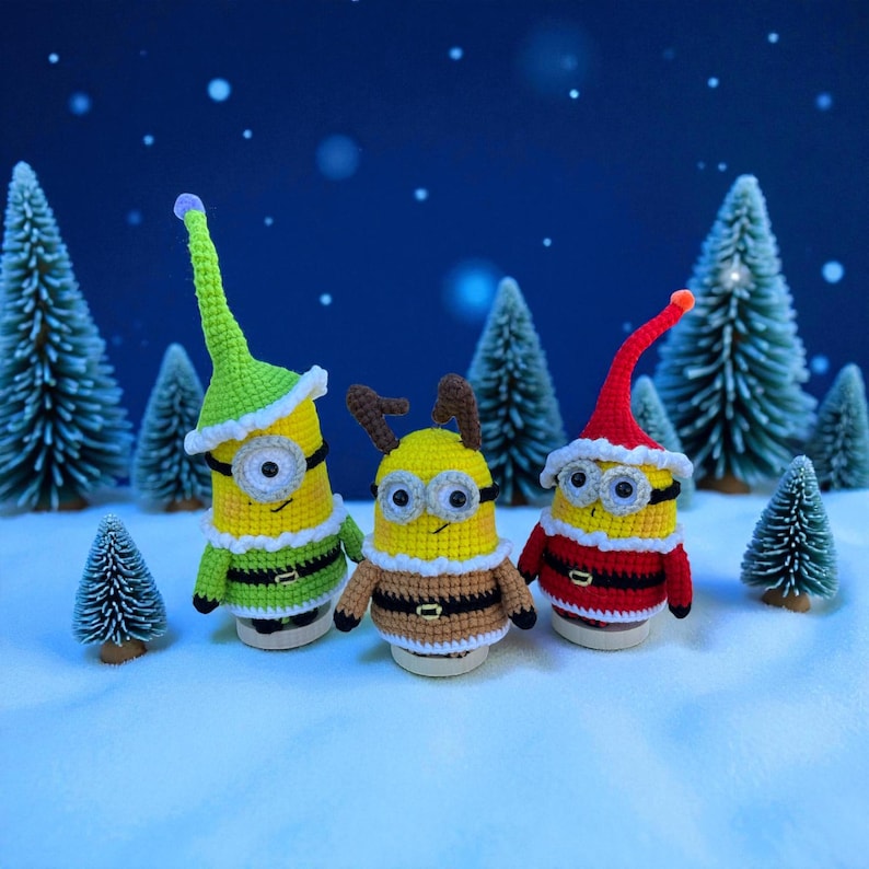 Może przedstawiać: Trzy szydełkowe figurki Minionk&oacute;w w świątecznych strojach. Jeden ma zielony kapelusz elfa, drugi rogi renifera, a trzeci czapkę Świętego Mikołaja. Stoją na zaśnieżonej powierzchni z miniaturowymi choinkami w tle.