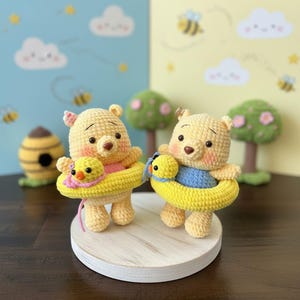 May include: Two crocheted yellow bear figures, each wearing a bright yellow swim ring and holding a small yellow duck. The bears have pink-tinted cheeks, dark eyes, and brown noses. One bear wears a pink shirt, and the other wears a blue shirt.