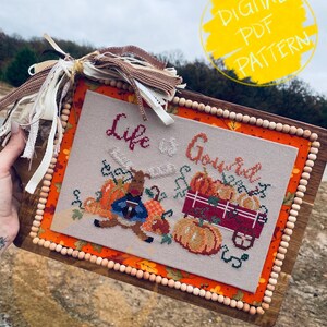 May include: Cross-stitch artwork with the phrase "Life is Gourd" and images of pumpkins and a wagon. The artwork is framed in a wooden frame with a beaded border and adorned with ribbons. A yellow circle with the text "DIGITAL PDF PATTERN" is in the background.