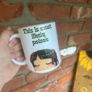 Könnte beinhalten: Weiße Keramiktasse mit dem Text "This is most likely poison" in Grün. Die Tasse zeigt eine Illustration im Anime-Stil eines Charakters mit schwarzen Haaren und roten Akzenten. Die Tasse hat einen weißen Griff.