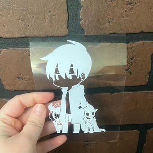 May include: A white vinyl decal featuring a stylized anime character with two animal companions. The character has spiky hair and is wearing a long coat. The decal is held in a hand against a brick wall.