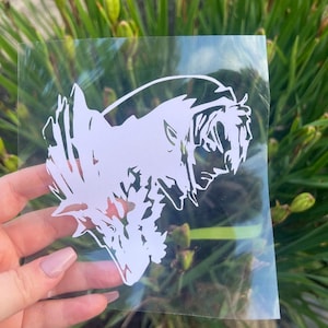 May include: A white vinyl decal featuring a stylized profile of a character with long hair and a wolf-like creature. The decal is held up against a background of green foliage, showcasing the intricate design and clean lines of the artwork.