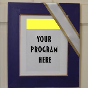 Puede incluir: Una impresión enmarcada con el texto "YOUR PROGRAM HERE". El marco es azul oscuro con un borde interior dorado. Una cinta blanca con borde dorado cruza diagonalmente el marco y un rectángulo amarillo está en la parte superior.