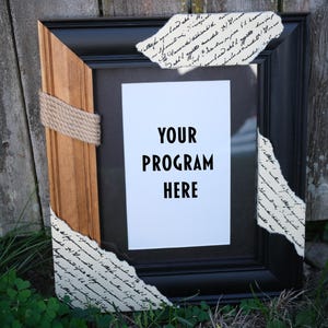 Puede incluir: Marco de fotos negro con un acento de madera y detalles de cuerda. El marco tiene un inserto blanco con el texto "YOUR PROGRAM HERE". Papel decorativo con escritura está adherido al marco.
