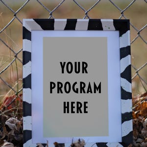 Puede incluir: Un marco de fotos rectangular con un patrón de rayas de cebra en blanco y negro. El marco contiene un fondo gris claro con las palabras "YOUR PROGRAM HERE" en texto negro. El marco descansa sobre hojas caídas.