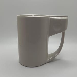 May include: A white ceramic mug with a unique design. The mug has a cylindrical body with a curved handle and a rectangular extension on the side. The mug is a solid, neutral color, and appears to be made of a smooth material.