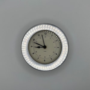May include: A silver round clock with a white face and black numbers. The clock has a silver rim with a textured design. The time is 12:00.