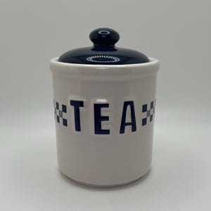 May include: White ceramic tea canister with a navy blue lid and knob. The word "TEA" is printed in navy blue block letters on the front, with navy blue and white checkered accents on each side of the word.