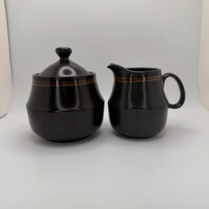May include: A matte black ceramic sugar bowl with a matching lid and a creamer pitcher. Both pieces feature a wide, horizontal band with a brown and black geometric pattern encircling the upper portion of the vessels, providing a subtle contrast.
