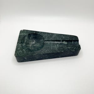 May include: A dark green marble ashtray with a rectangular shape and a circular indentation for holding cigarettes. The ashtray has a groove for resting cigarettes and a smooth, polished surface. The ashtray is a stylish accessory for smokers.
